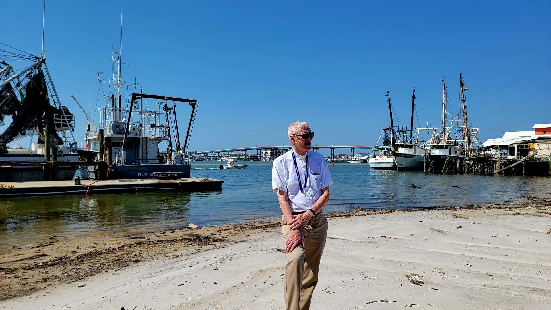 Man standing on a beach with fishing boats docked in the background on a sunny day.