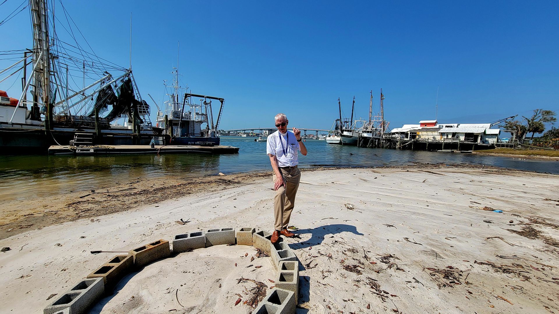 Man stands on a sandy beach with boats docked behind him on a sunny day.