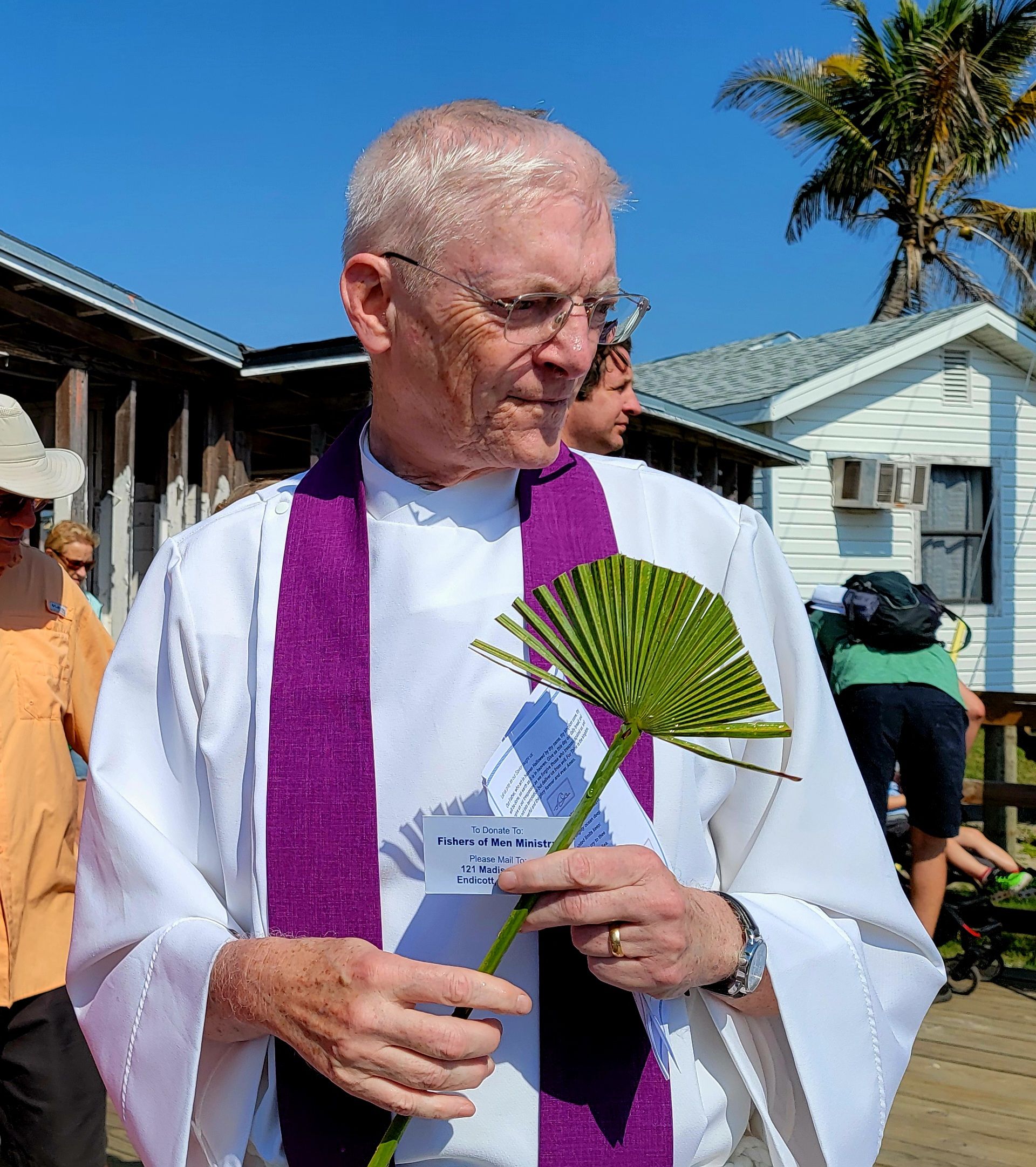 Elderly man in white robe with purple stole holding palm frond outdoors.