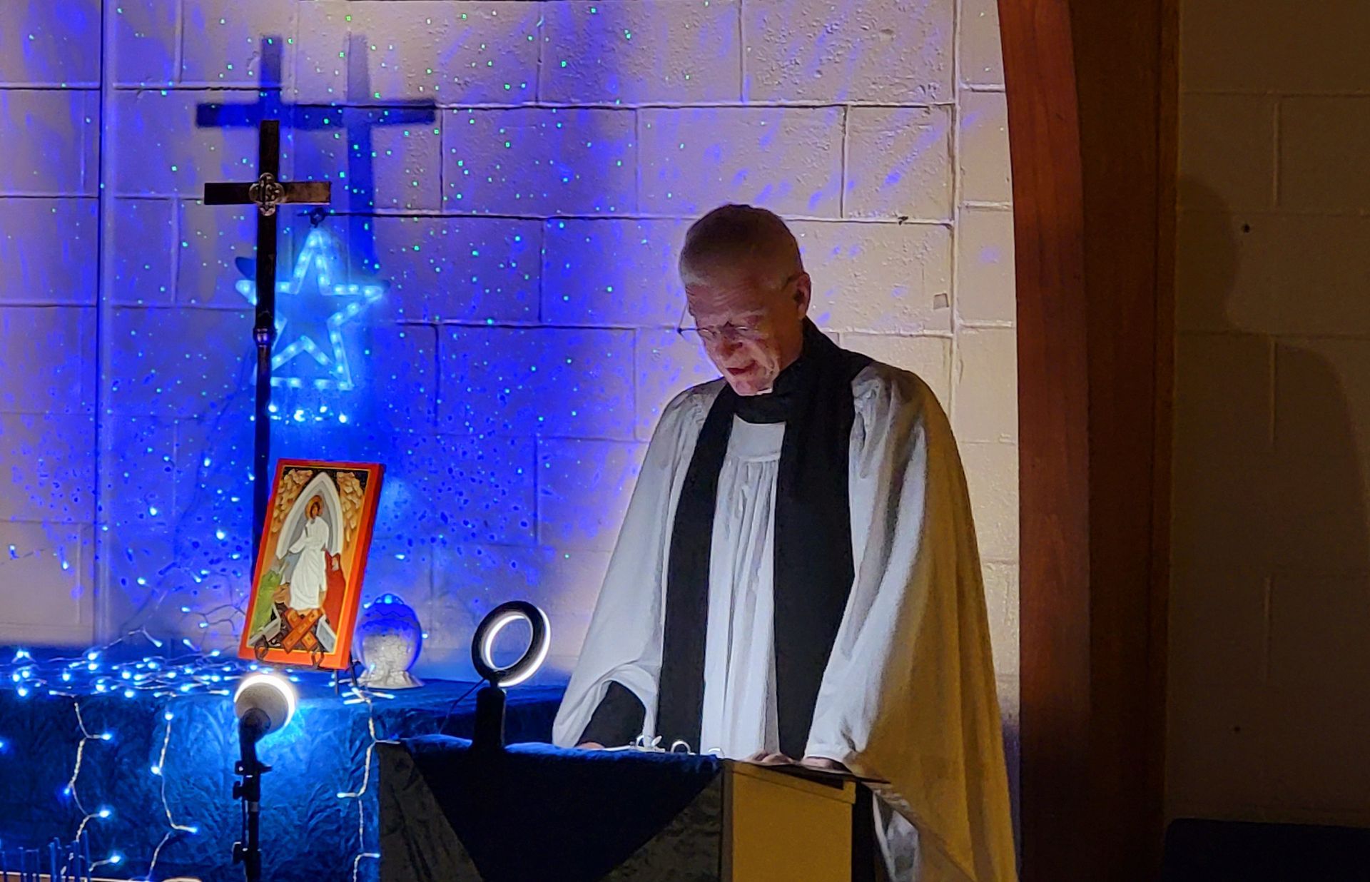 Clergyman in white robe reads at a podium, cross and blue lights in the background.