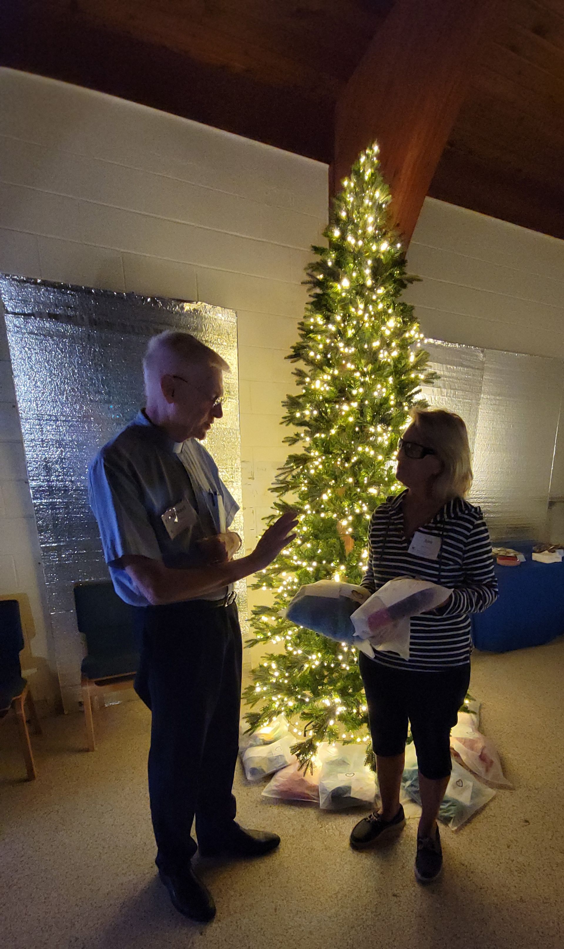 Man and woman beside lit Christmas tree, indoors. Man gestures; woman holds gifts.
