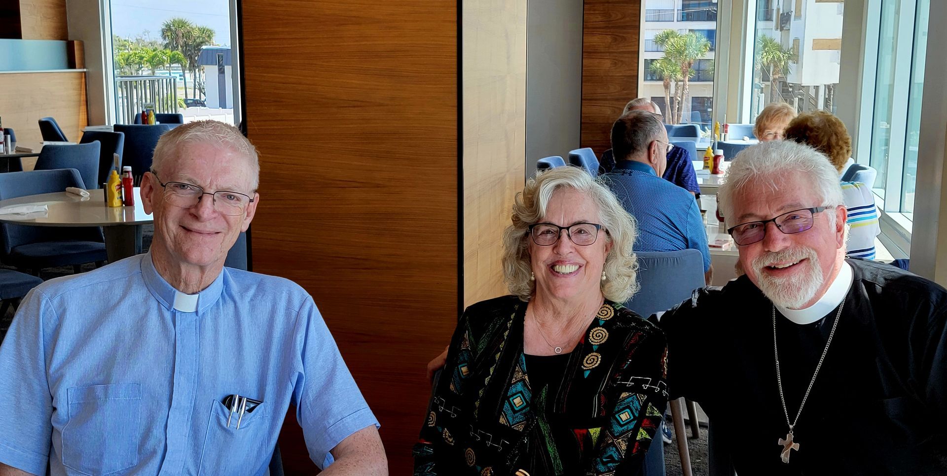 Three people smiling at a table in a restaurant, two men in clerical collars.