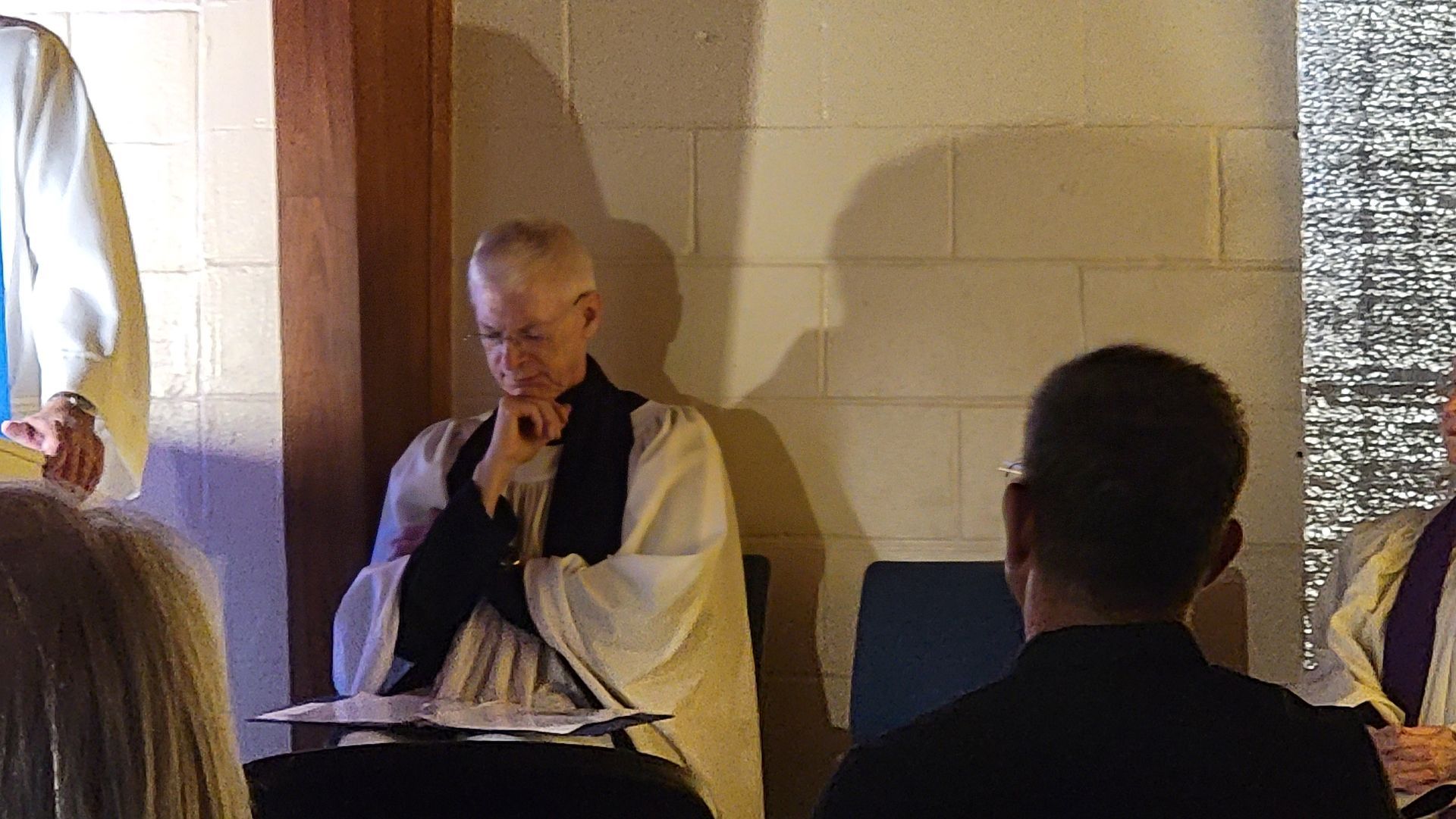 A clergy person in white and black robes sits thoughtfully, hands together, inside a church.