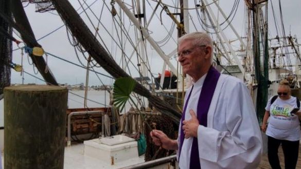 A priest in white and purple robes blesses a boat on a cloudy day at a dock; a woman watches.