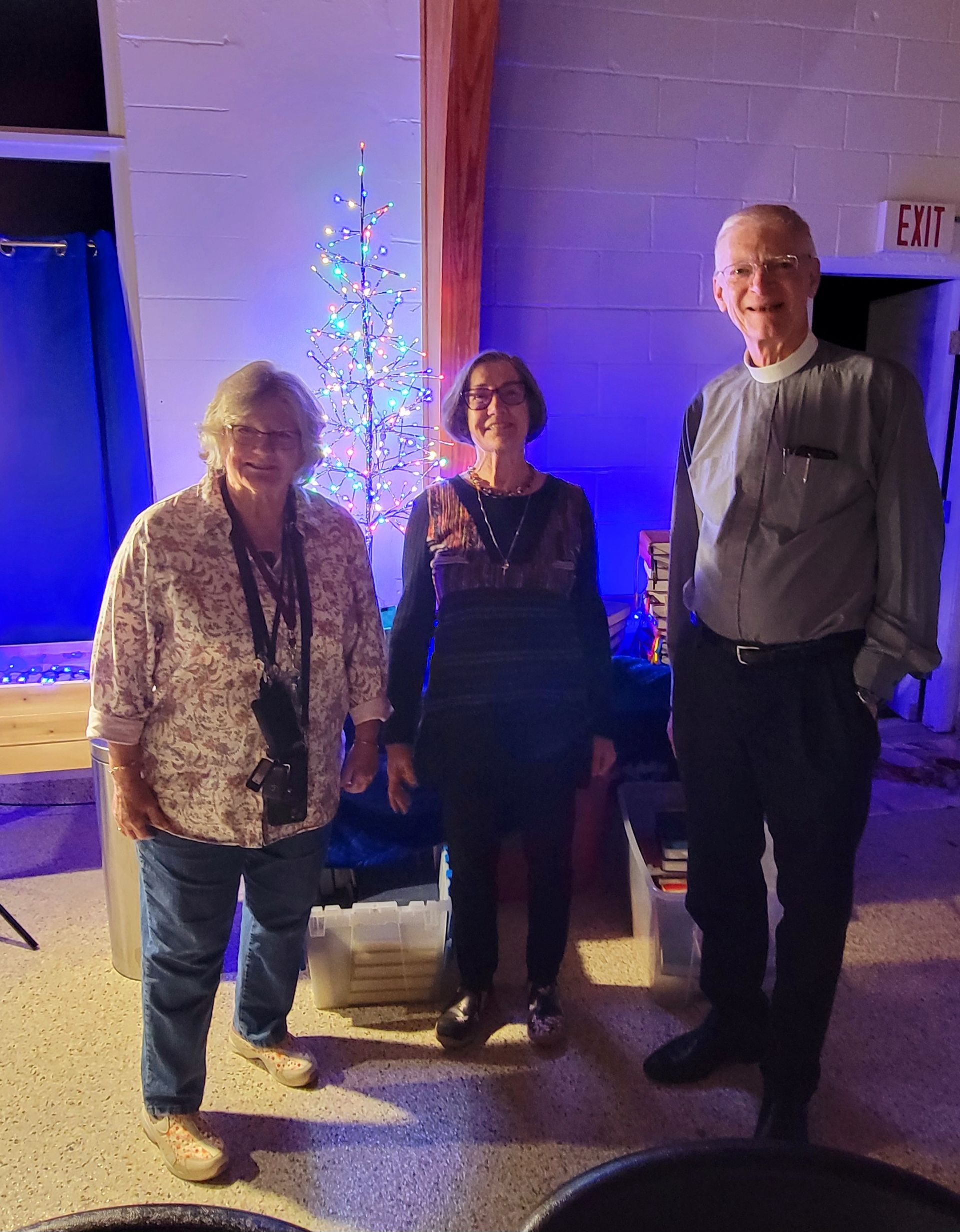 Three people stand indoors; woman in a patterned shirt, woman in a dress, and man in a button-down shirt