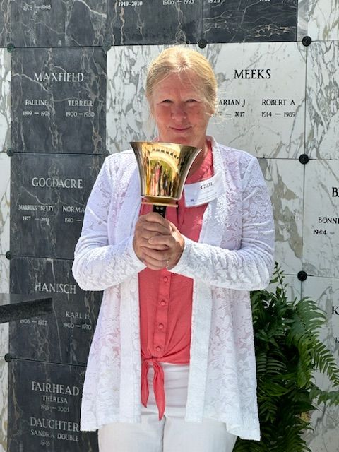 Woman in white cardigan holds a golden bell in front of a mausoleum.