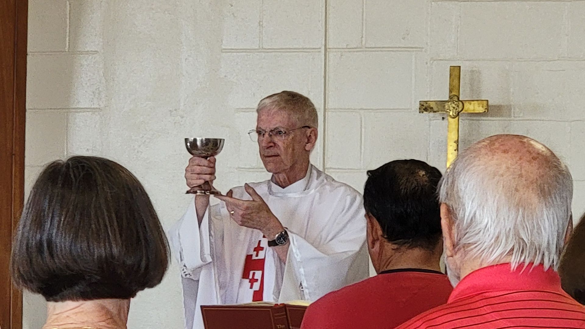 Priest holds up chalice during a church service. People watch in a white-walled room with a cross.