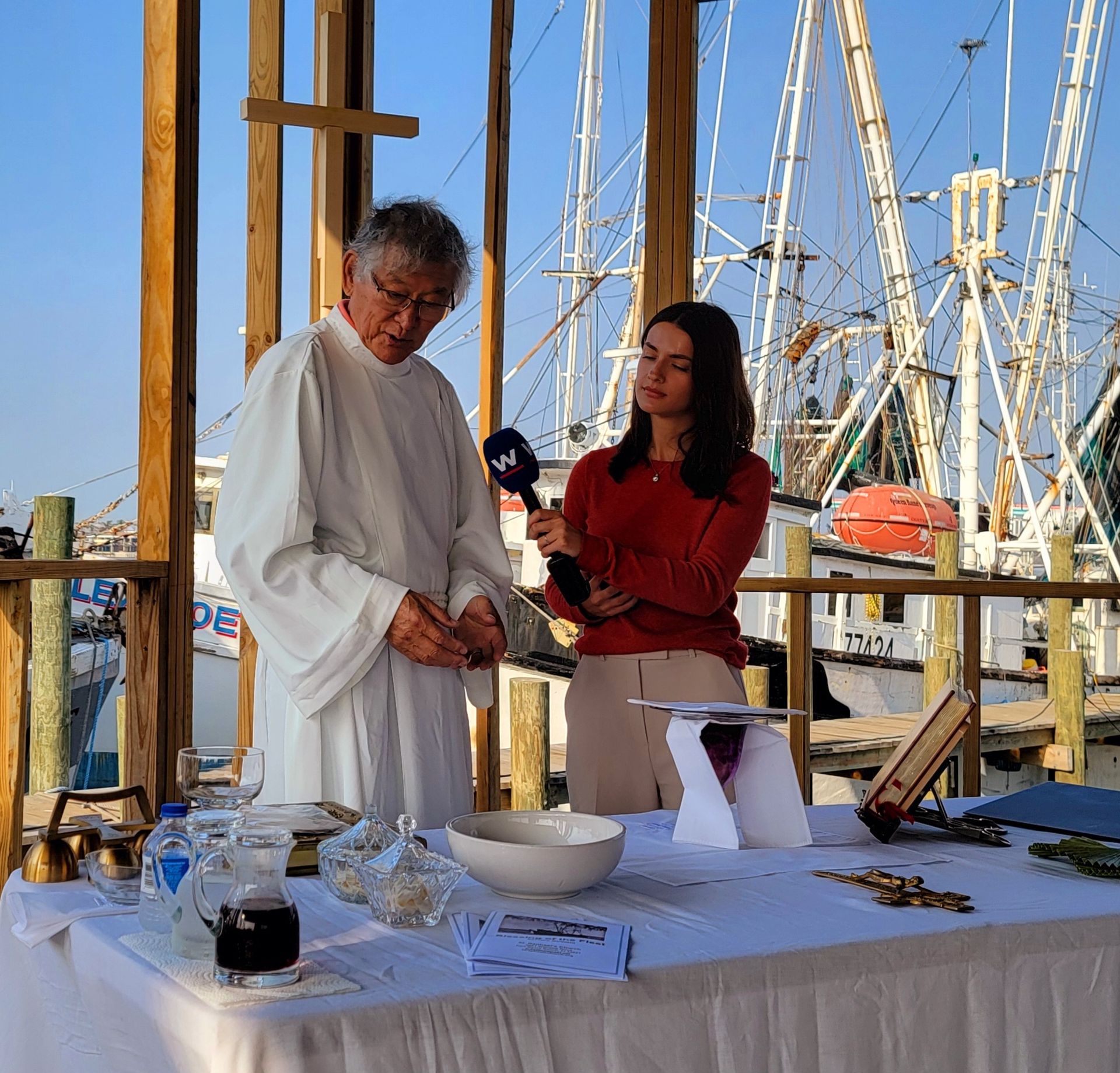 A priest being interviewed at an outdoor waterfront ceremony with a reporter.  Boats and blue sky in the background.