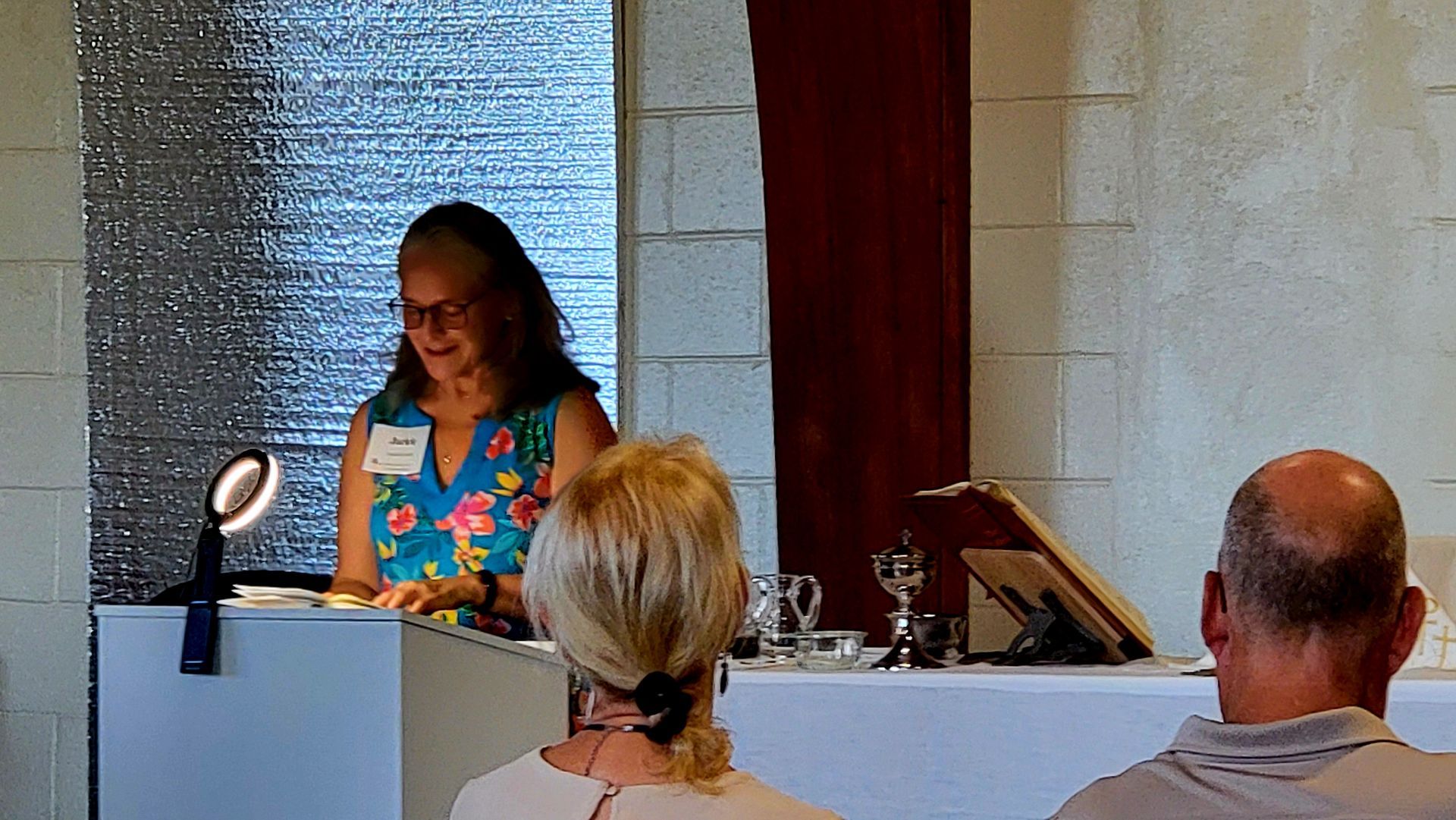 Woman speaking at a podium, floral top, silver background, two audience members.