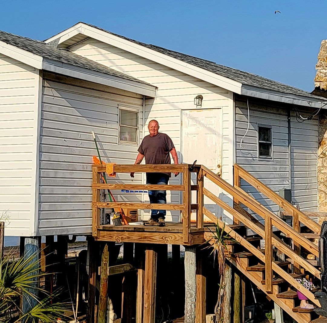 Man on a wooden porch of a stilted white house. Sunny day, blue sky.