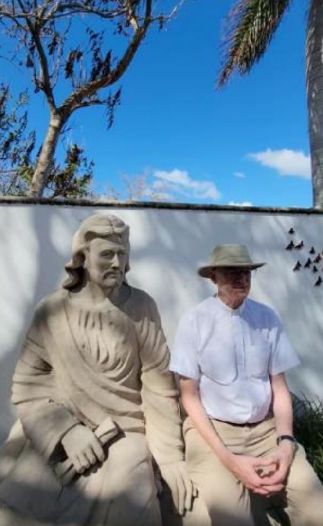 Man in a hat sits next to a statue in front of a white wall under a bright blue sky.