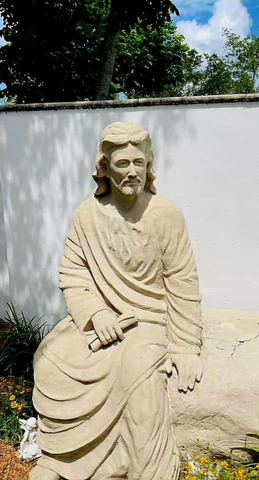 Statue of Jesus seated on a rock, holding a scroll, against a white wall and blue sky.