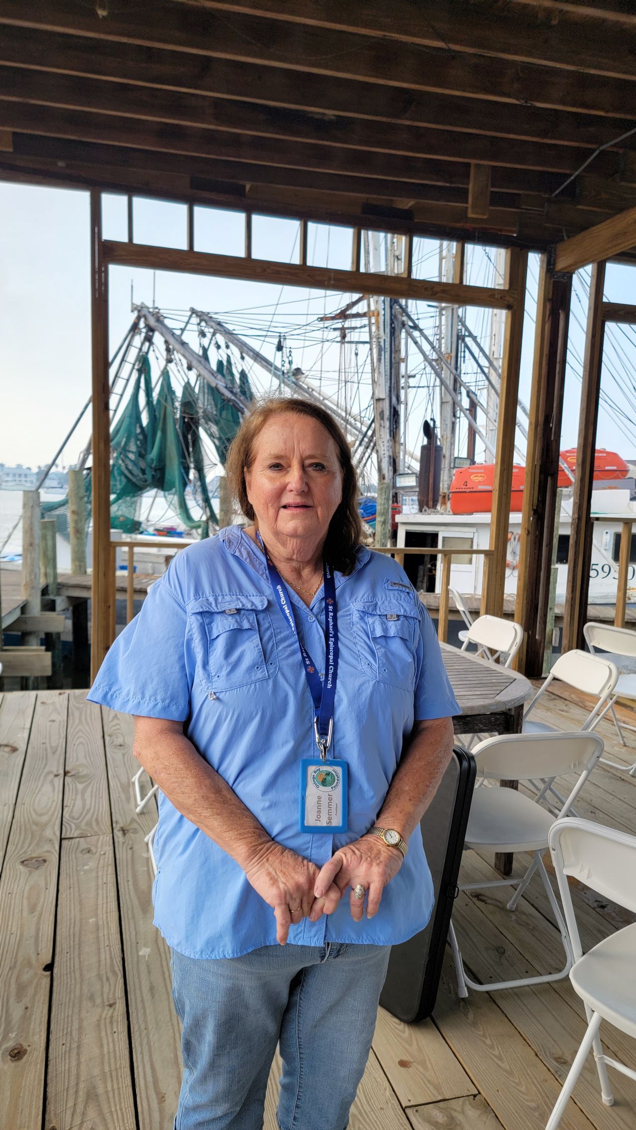 Woman in blue shirt, jeans, standing on a wooden dock with a boat in the background.