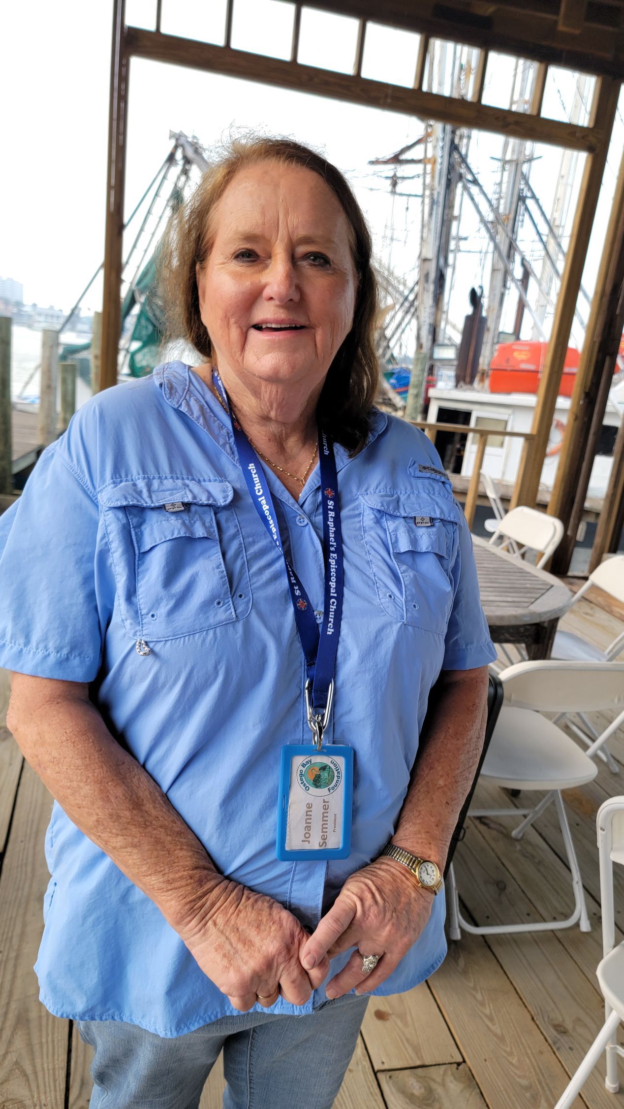 Woman in blue shirt stands on wooden deck; shrimp boats in background.