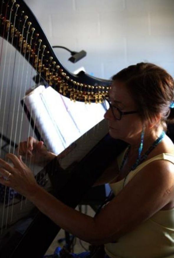 Woman playing a large black harp, wearing glasses and a turquoise necklace.
