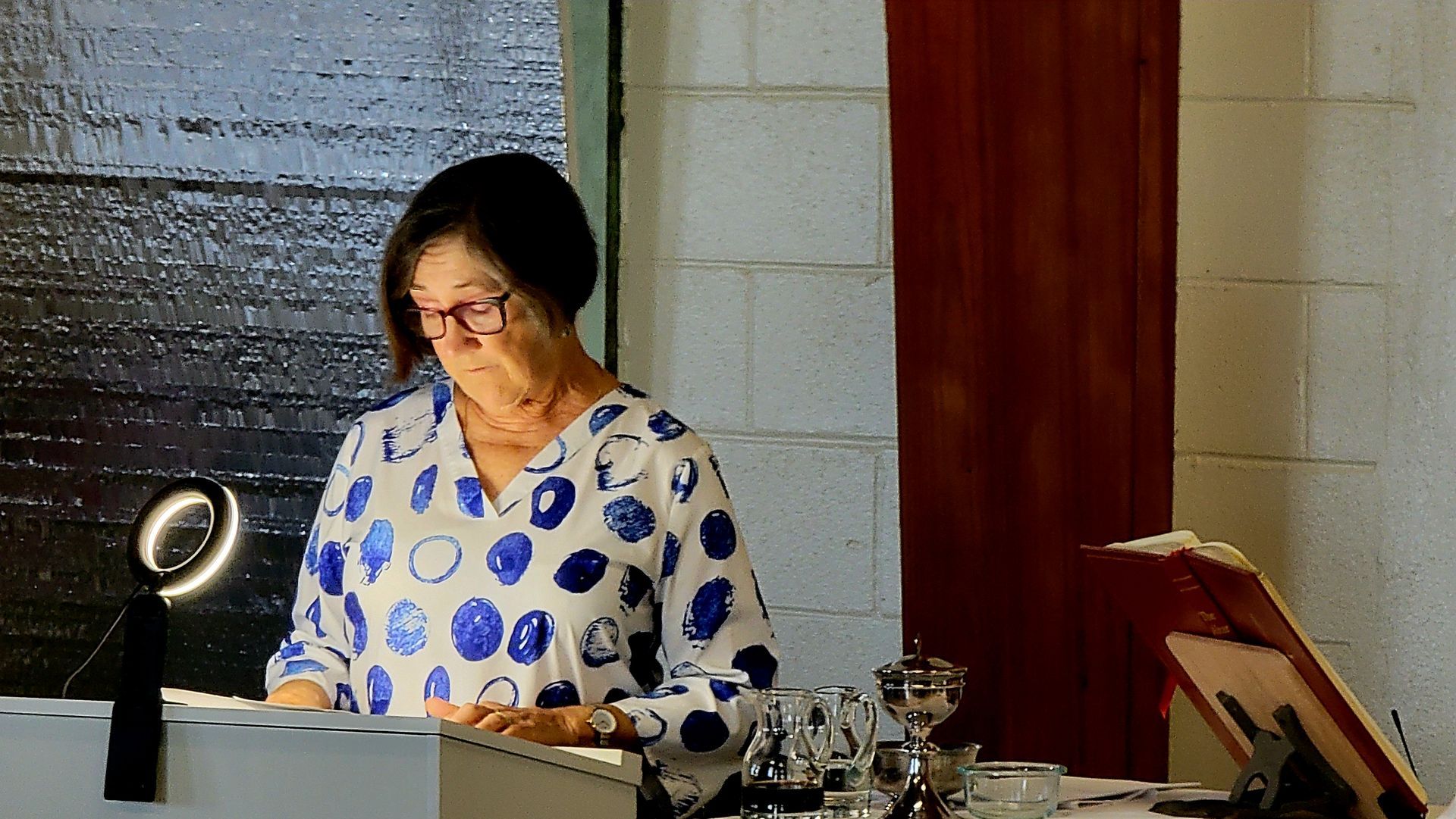 Woman in patterned shirt plays a keyboard, lit by a ring light, in a room with a wooden door.