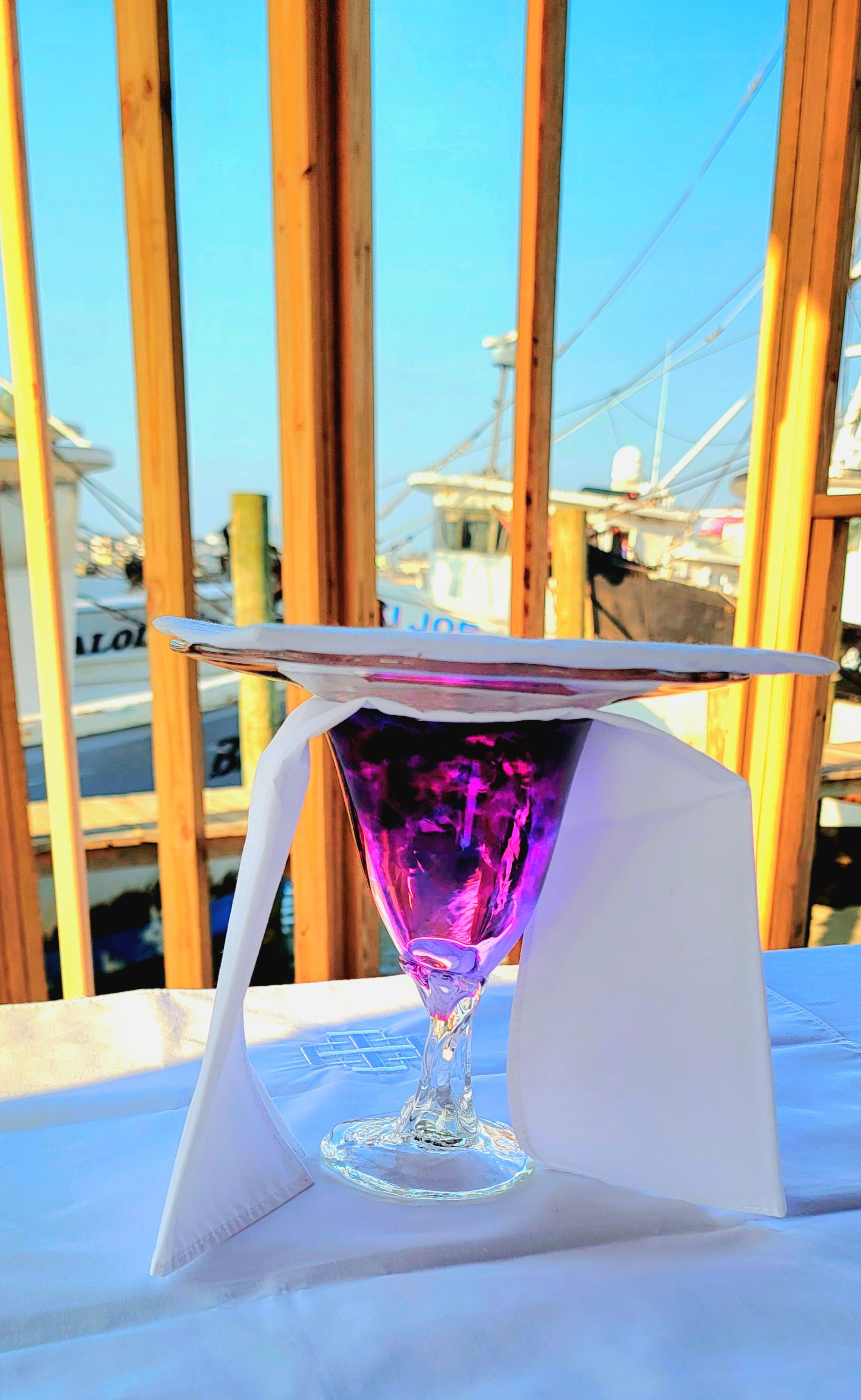 Purple cocktail in a glass, on a white napkin, on a table. Boats in the background.