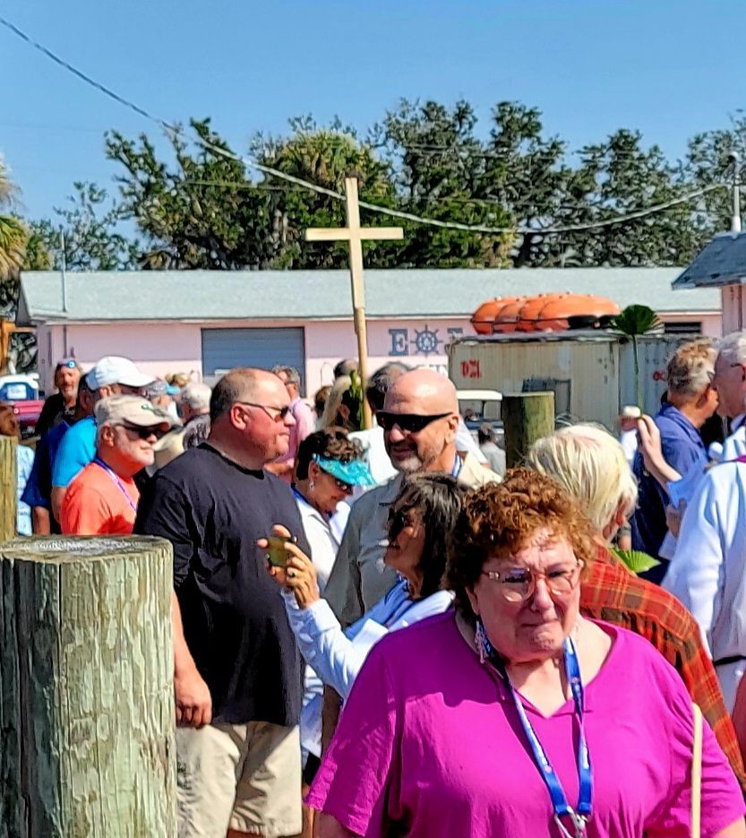 People gathered outdoors; a cross is held aloft. Pink building and trees in background.