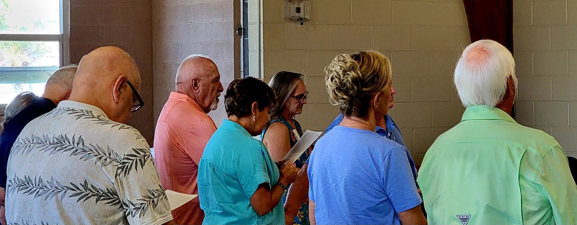 A group of people stand indoors; some look towards something unseen, various skin tones and clothing colors.