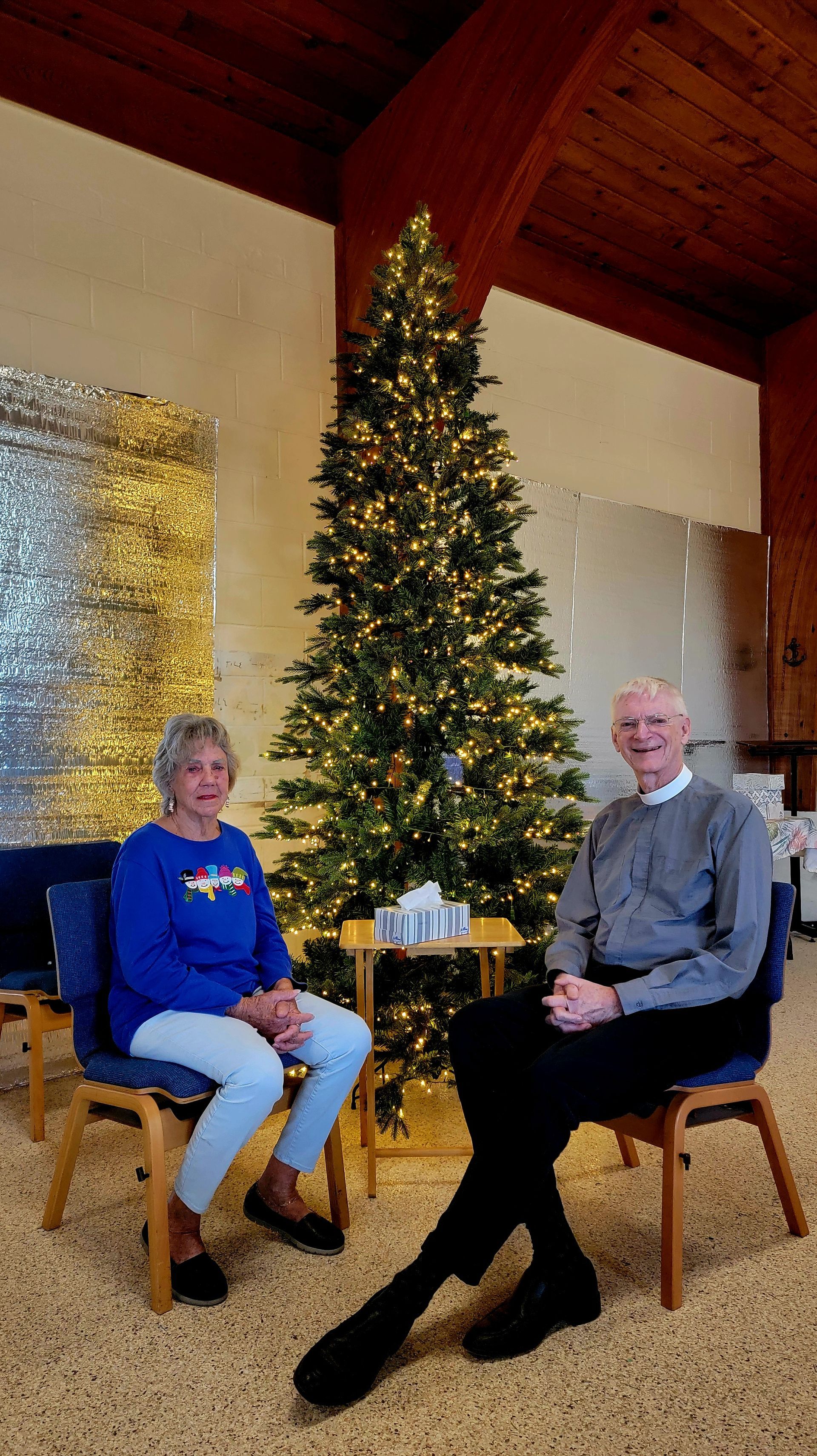 Woman and man sit beside a lit Christmas tree in a church.