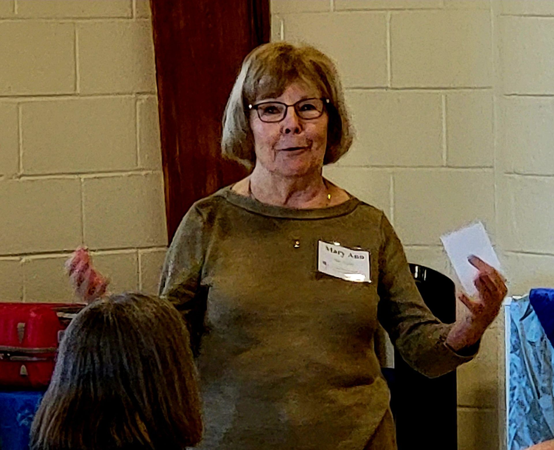 Woman with glasses and a name tag speaking, holding a paper, gesturing. Brick wall background.