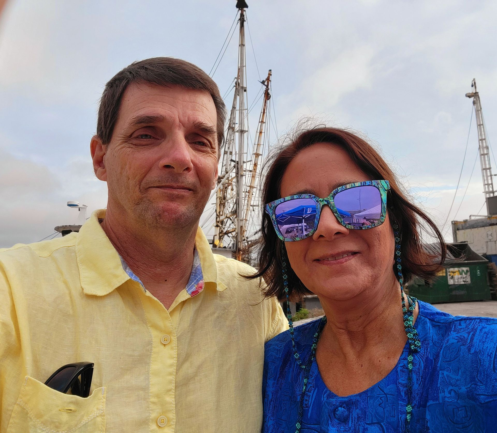 Couple smiling in front of a boat. Man in yellow shirt, woman in blue. Cloudy sky.
