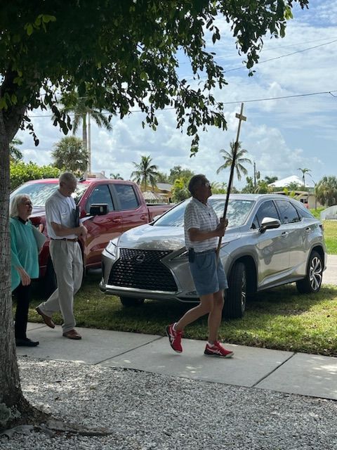 Group of people walking on sidewalk, man holding staff, cars parked nearby, sunny day.