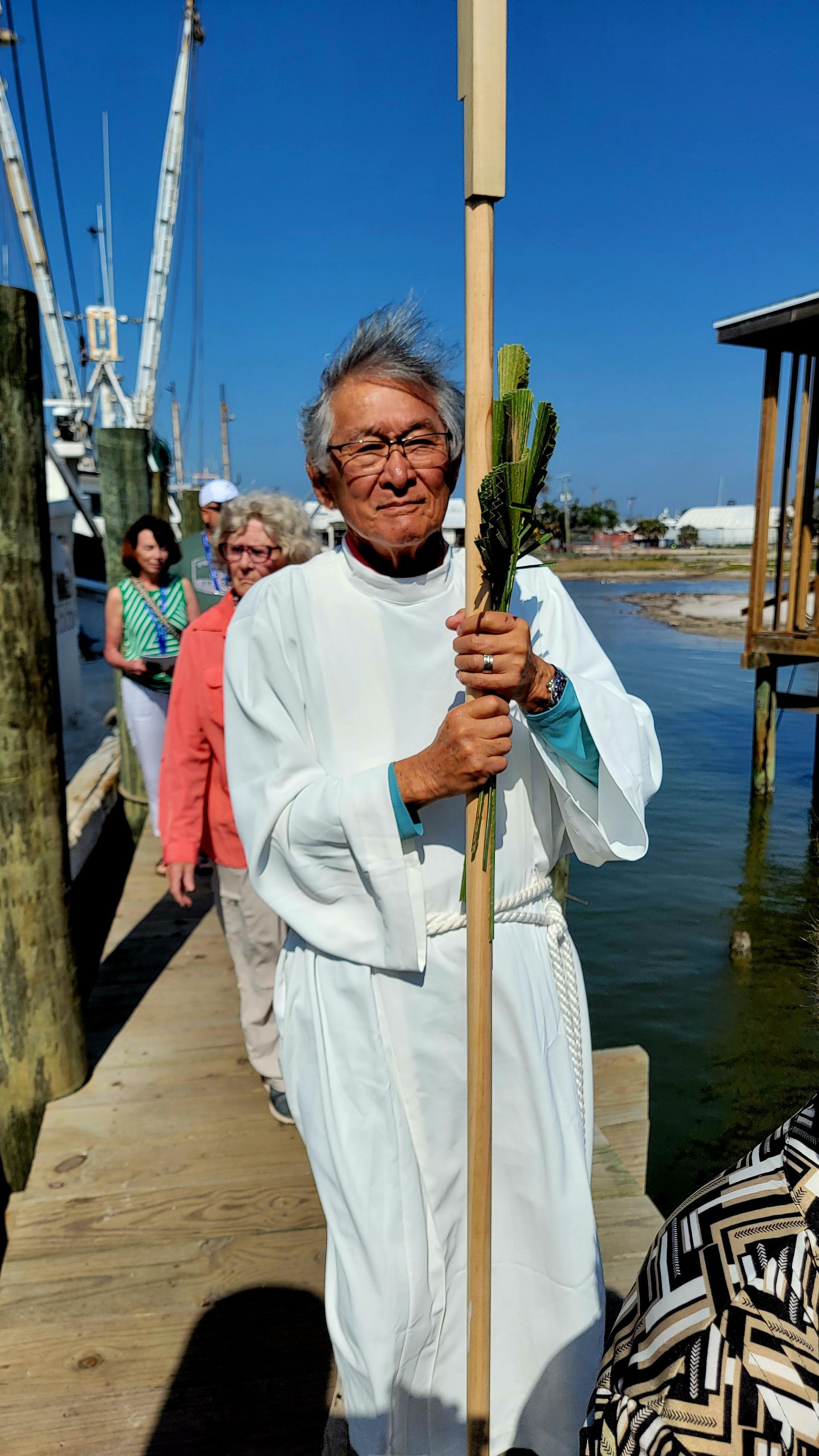 Man in white robes holds a staff with greenery, leading a procession on a wooden pier near water.