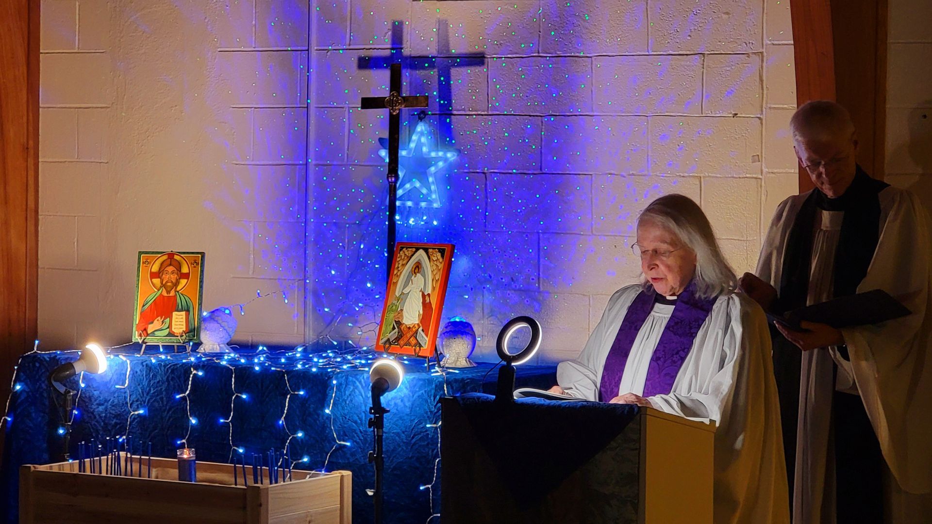 Clergy at altar with candles, icons, and a cross; blue lights illuminate.