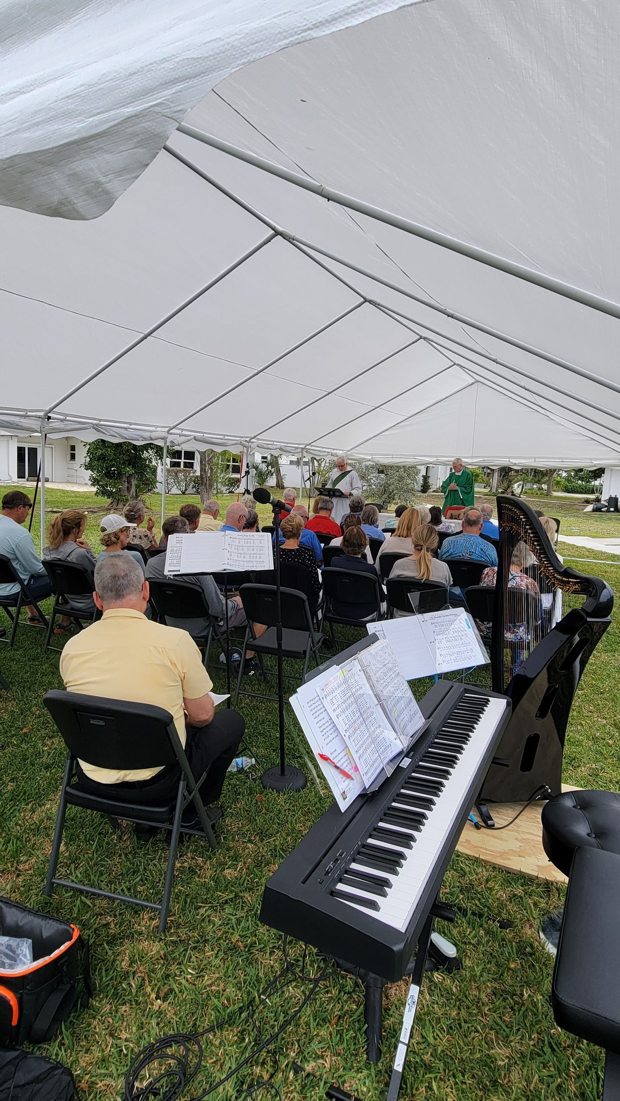 Musicians and audience under a white tent on grass. Keyboard in foreground, sheet music.