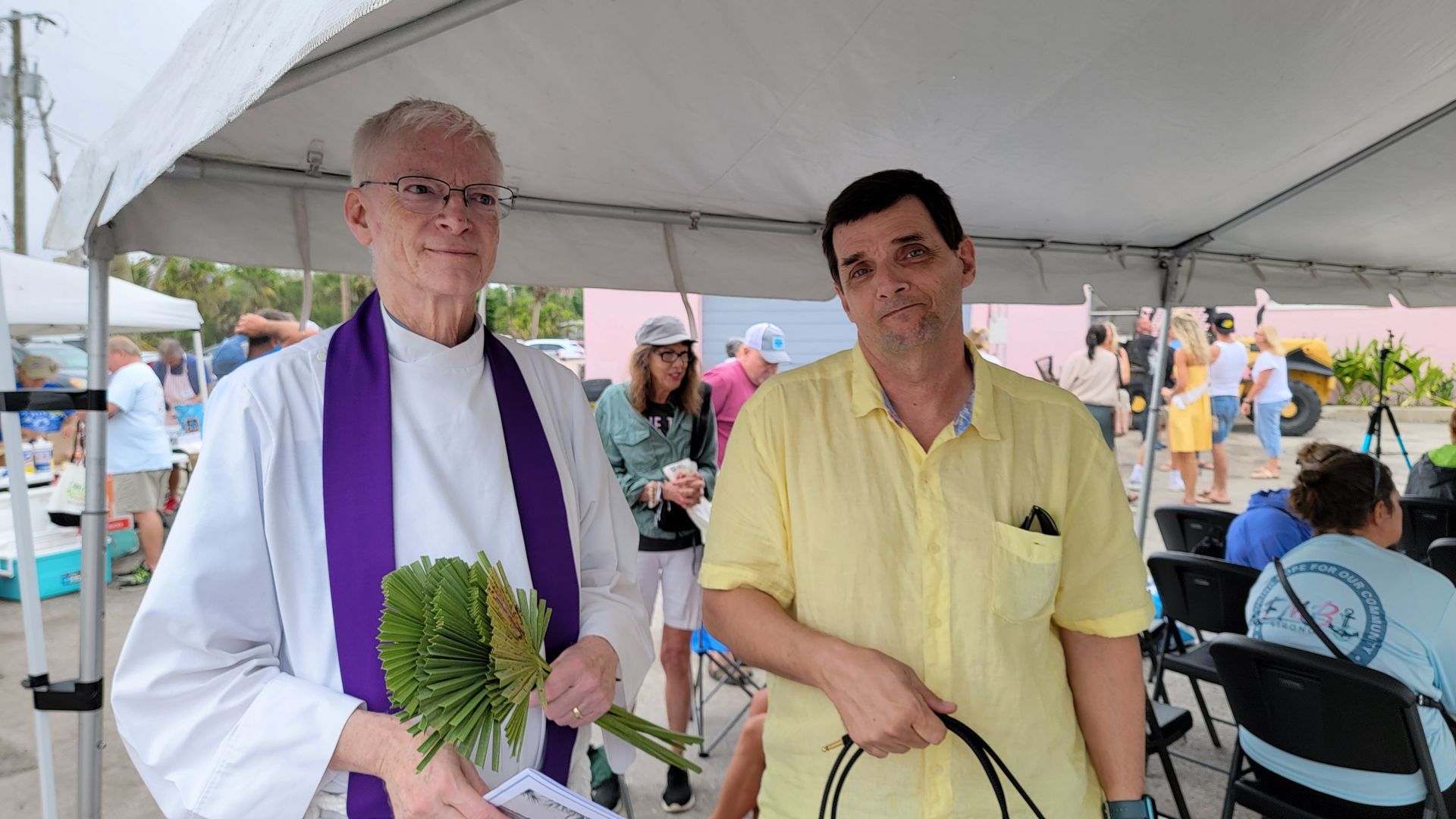 Man in clerical vestments with palm fronds next to a man in yellow shirt outdoors under a tent.