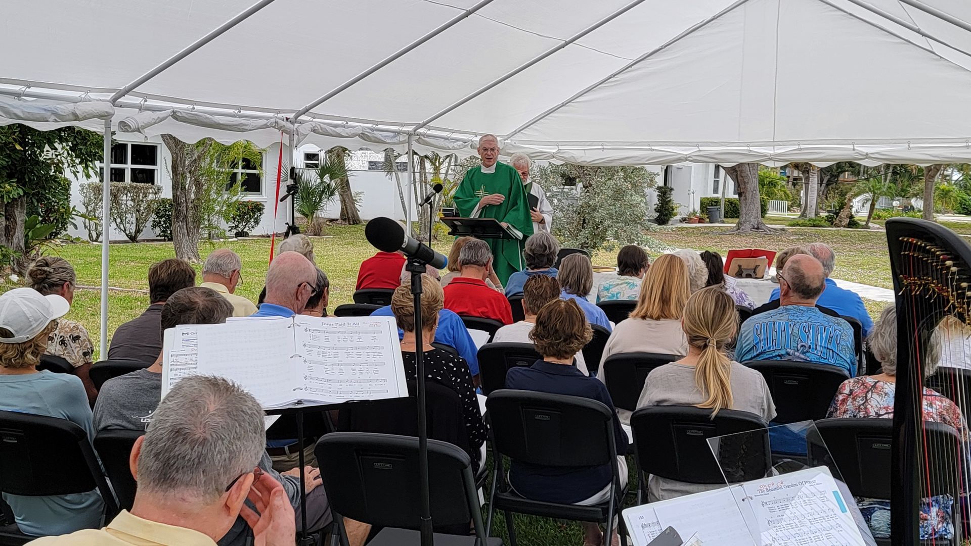 Church service under a white tent. A priest speaks to seated people, with musical instruments present.