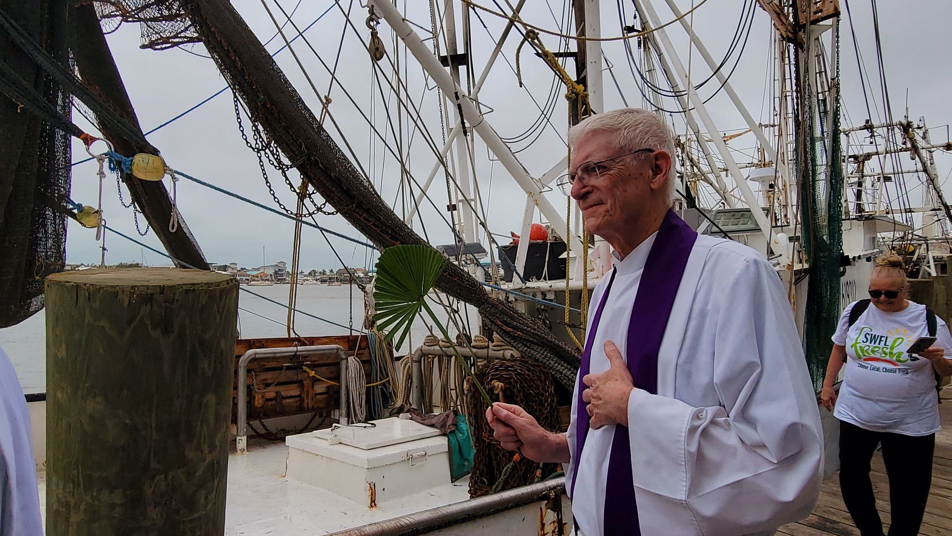 A priest blesses a shrimp boat at a dock. He wears white and purple robes. Gray sky, water, and ropes.