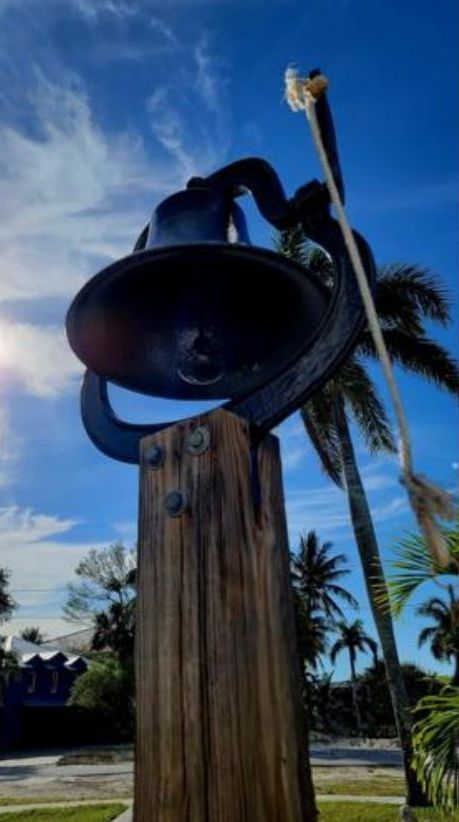 Large, dark bell mounted on a weathered wooden post under a blue sky with palm trees.