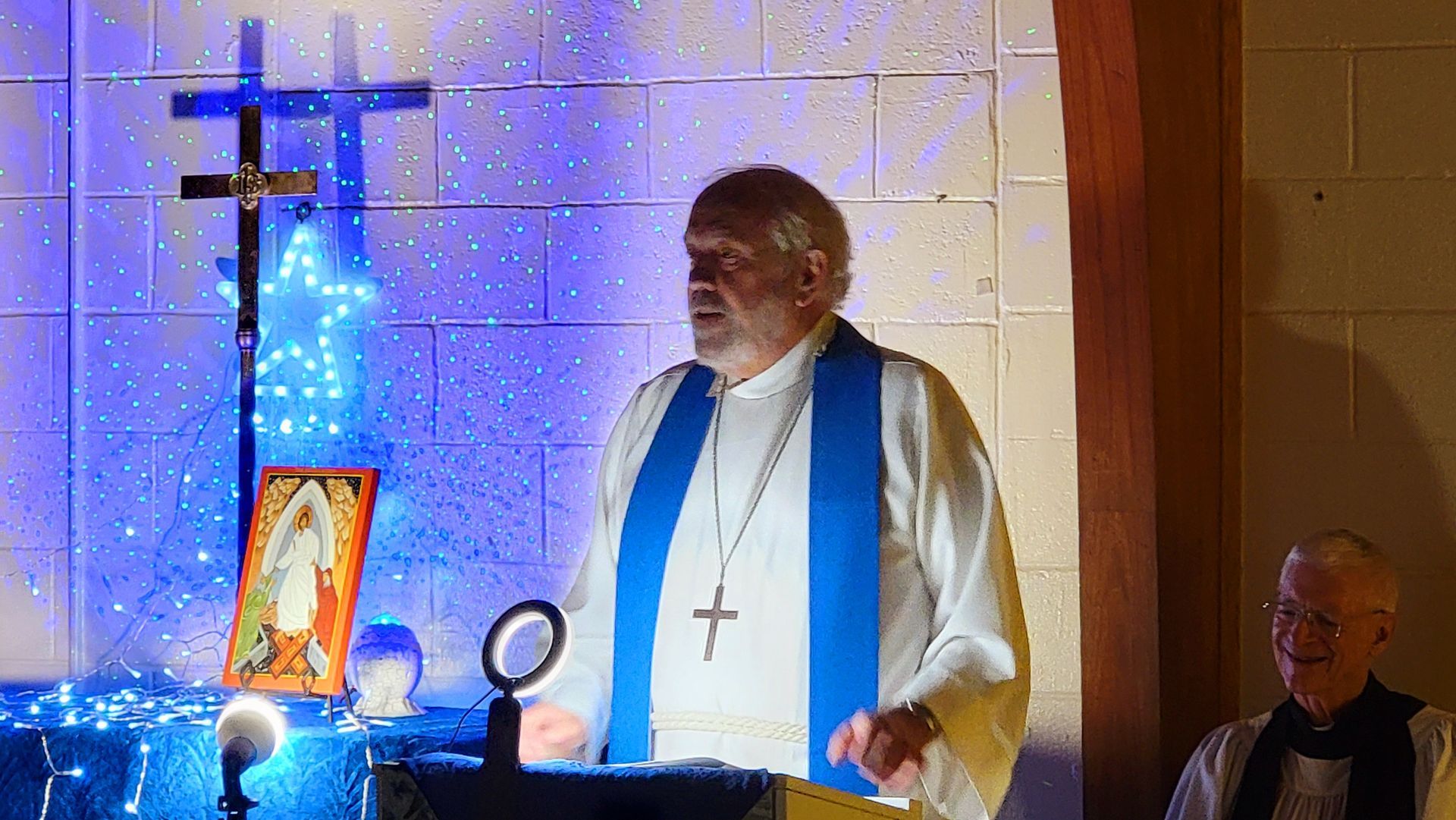 Man in clerical collar speaking at a pulpit; blue lighting, crosses, icon in the background.