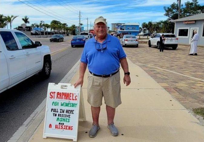 Man holding sign for St. Raphael's Ash Wednesday. Cars and buildings in background.