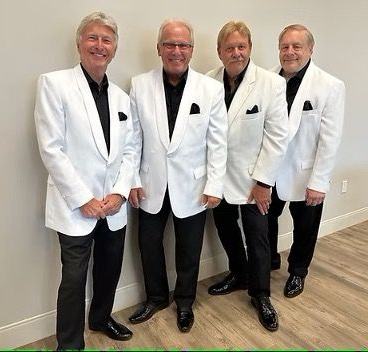 Four men in white jackets and black pants pose together against a light wall.