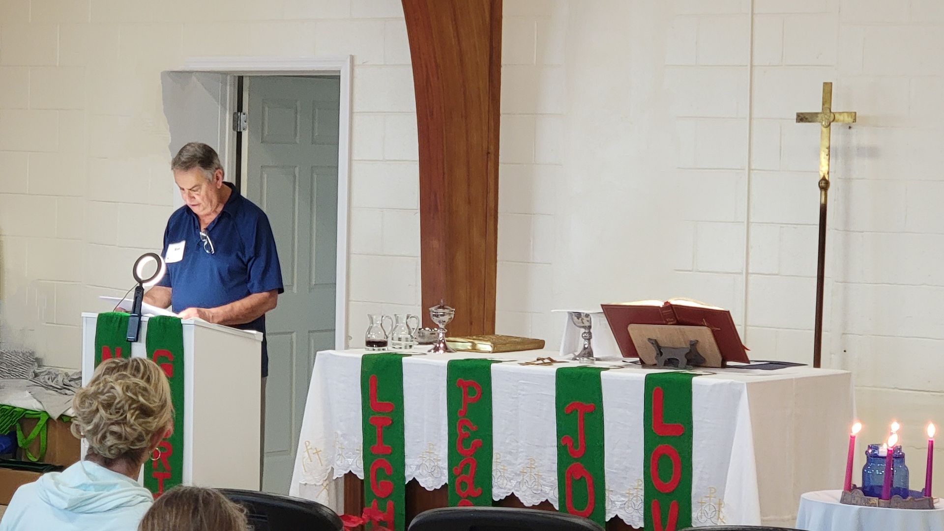 Man reading from a podium in a church, with a cross, candles, and people in the congregation.