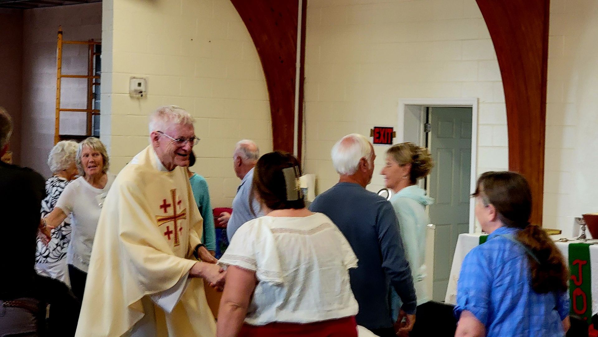 Priest greeting churchgoers inside a church. Smiles and handshakes. Light-colored walls, and a green banner.