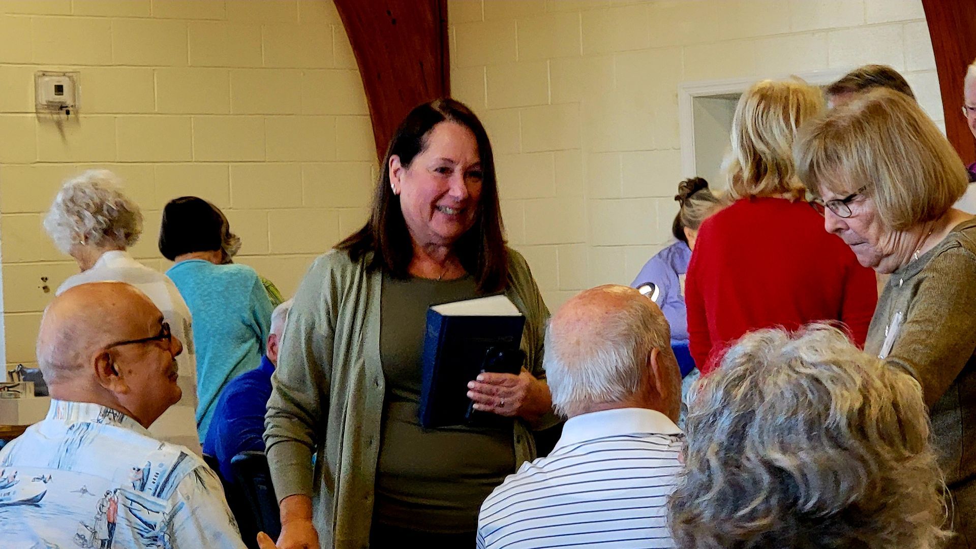Woman in green top smiles and talks to people in a room. People surround her, some seated.