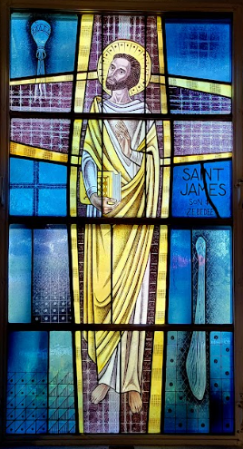 Stained glass window of Saint James, holding book, gold robe, halo, surrounded by blue and gold.