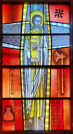 Stained glass window of Saint Matthew holding book and axe, surrounded by symbols and text.