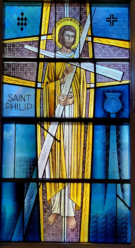 Stained-glass window of Saint Philip holding a cross. Yellow robe, halo, and beard. Blue background.