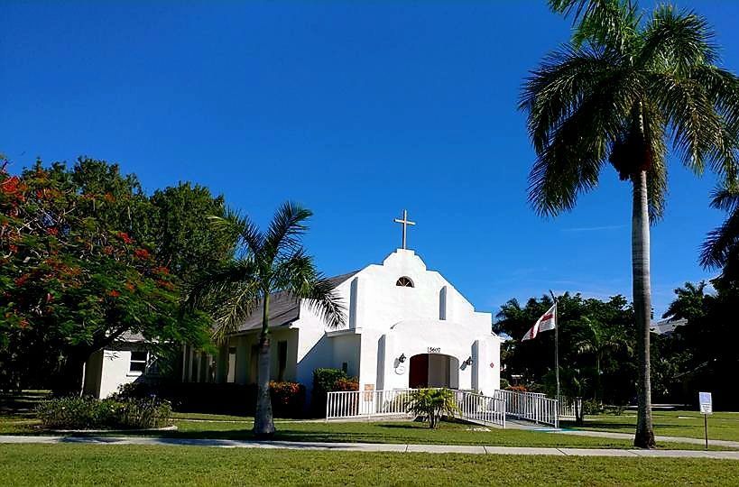 White church building with cross on a clear, blue sky day, flanked by palm trees and green lawn.