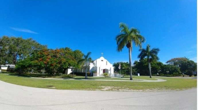White church building with a cross on top, surrounded by palm trees and green grass on a bright, sunny day.