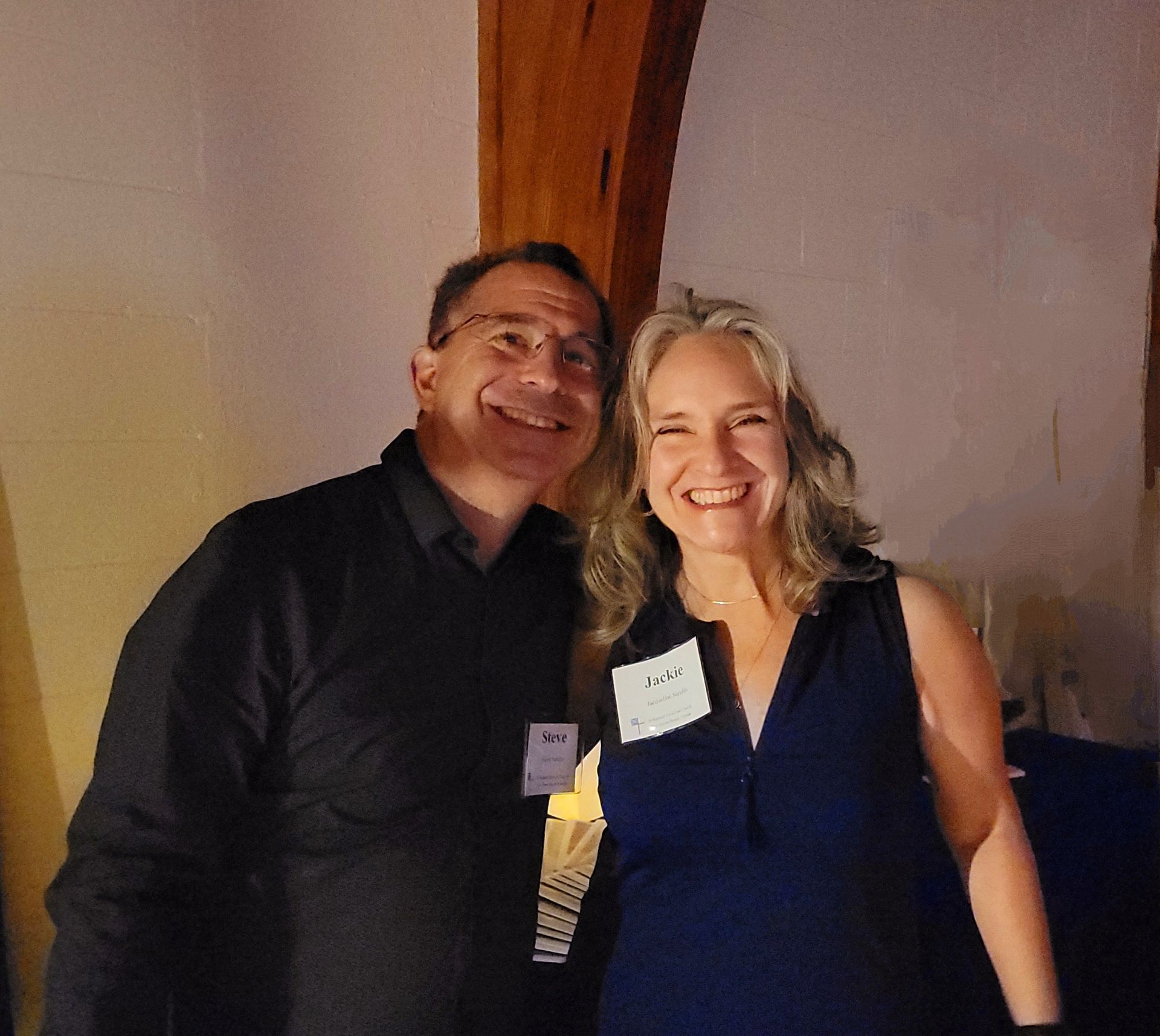 Two smiling people, a man and a woman, posing together. They are indoors.