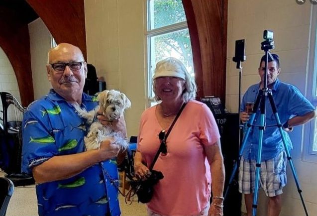 Man holding small dog, woman with camera, and man with tripod in a room.