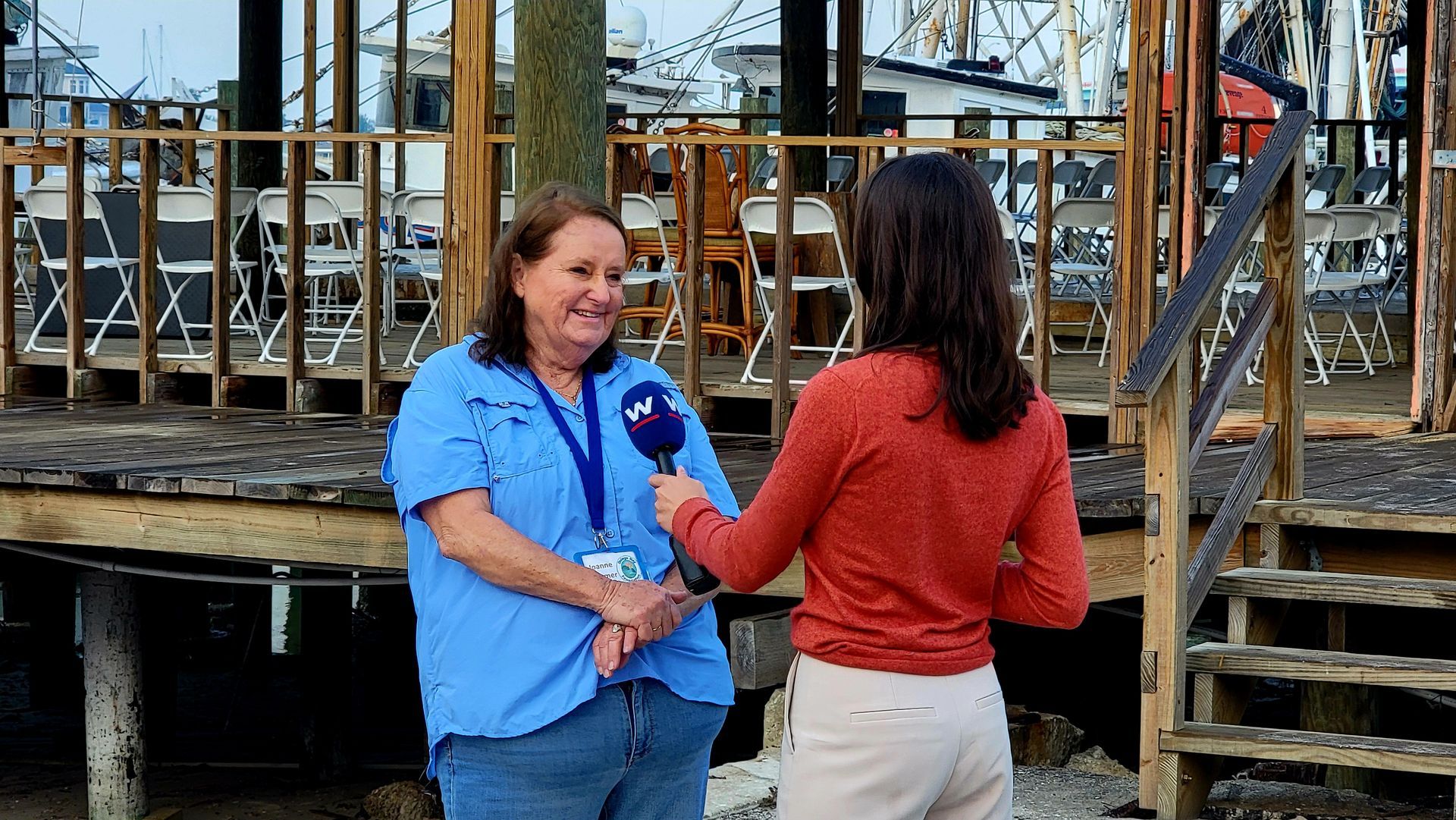 Woman in blue shirt being interviewed outdoors, smiles at reporter holding microphone. Deck, boats in background.