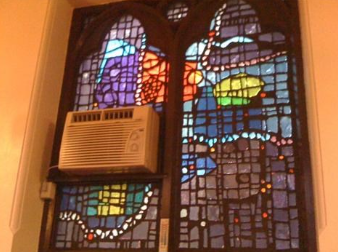 Stained-glass window with an air conditioner installed in it; colorful design with purple, orange, blue, and green hues.