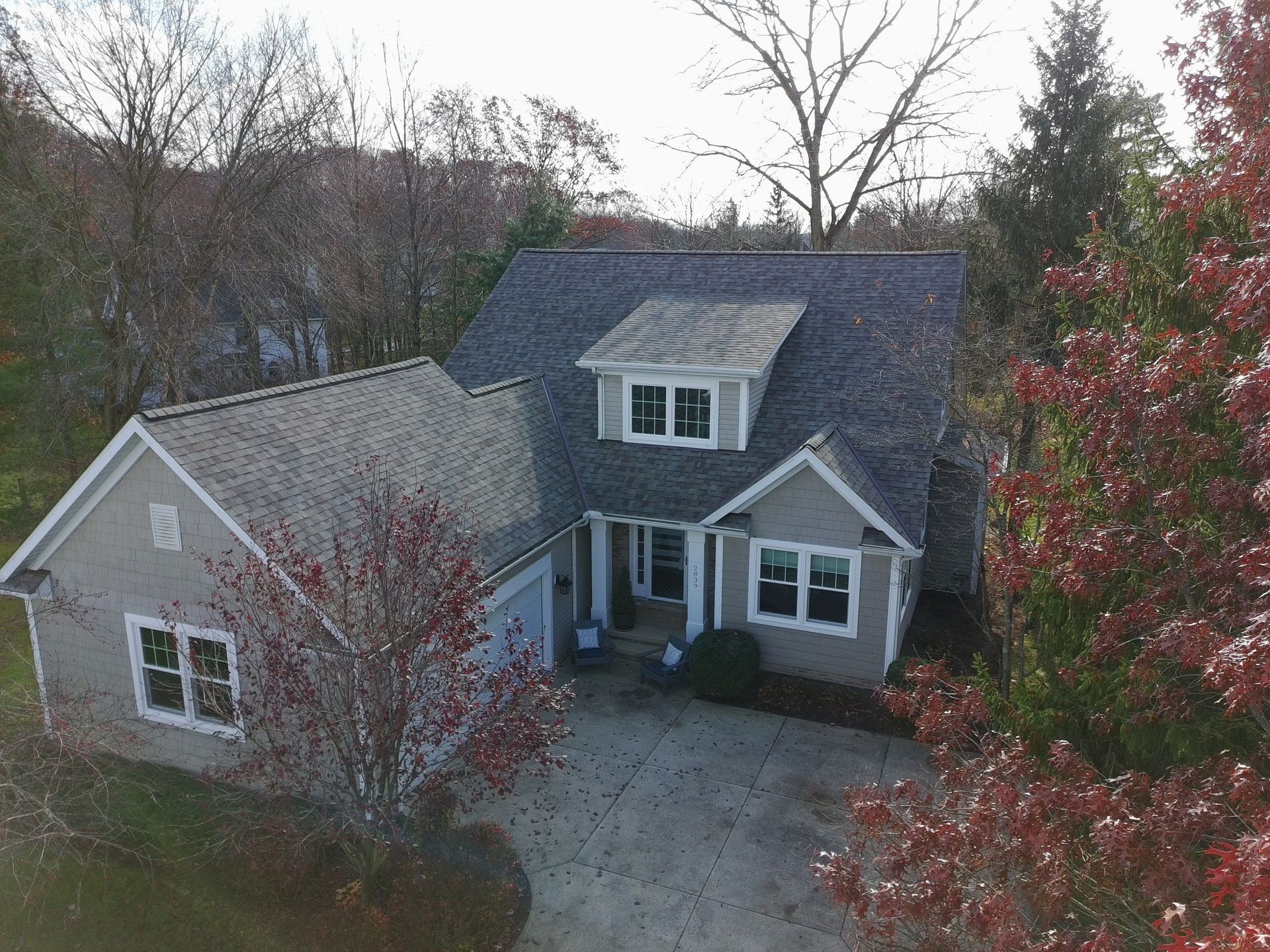 An aerial view of a gray-sided suburban home with a dark shingled roof, set among trees in an autumnal landscape.